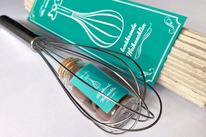 Weihnachtsmailing mit Schneebesen und kleinem Glas voller Schokodrops, verpackt im türkisfarbenen Design mit Aufschrift ‚berührende Weihnachten‘.