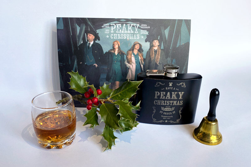 Weihnachtsmailing im ‚Peaky Blinders‘-Stil mit Karte, Flachmann, Whiskeyglas, Mistelzweig und Glocke.